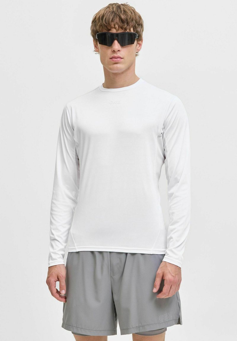 Vit långärmad skjorta med en slät textur, parat med grå shorts. Modellen bär svarta solglasögon. Enkelt, atletiskt mönster, inga mönster.