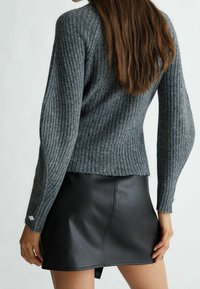Pull gris à côtes avec des manches ajustées, associé à une mini jupe en cuir noire. Détails notables : un accent en bijoux sur la manche du pull.