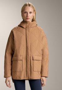 Jachetă matlasată cu glugă, de culoare brown deschis, cu două buzunare frontale și o croială lejeră. Textură netedă cu un luciu subtil.