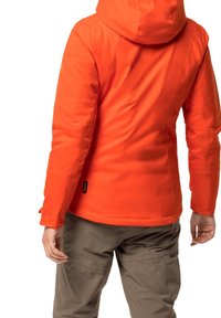 Oranžová zateplená bunda s kapucí, hladkou texturou, švy a značkou s logem u dolního lemu. Kombinována s béžovými kalhotami.