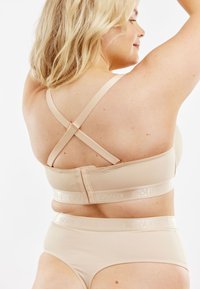 Beige bh en slipset van glad, rekbaar materiaal. De bh heeft verstelbare kruislings geplaatste bandjes en een logoband; de shorts hebben een brede elastische tailleband.