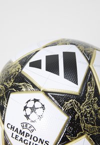 UEFA Champions League fotball, med en hvit overflate med svarte og gull aksenter, stjernedesign og teksturerte paneler. Adidas-logoen er tydelig synlig.