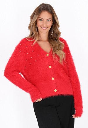 Rode fluffy cardigan met V-hals, voorzien van gouden knopen en verspreide rhinestones op de linker schouder, gedragen over zwarte broek.