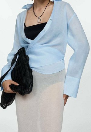 Person iført en lyseblå omslagsbluse over sort top, beige nederdel, sort halskæde og holder en sort flettet håndtaske.