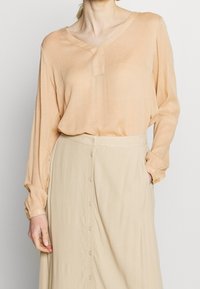 Blouse de couleur pêche à manches longues et col en V, associée à une jupe beige avec une rangée centrale de boutons. Tissu lisse et léger.