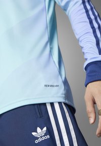 Lys blå atletisk skjorte med gradientmønster, fremhævet af marineblå striber på ærmerne. Har "AEROREADY" logo tæt på kanten. Parret med marineblå shorts.