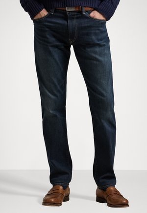 Man draagt donkerblauwe spijkerbroek, bruine leren riem, bruine loafers en een donkerblauwe trui met handen in de zakken op een witte achtergrond.
