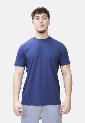 Giovane uomo in piedi, rivolto verso di noi, che indossa una t-shirt a maniche corte blu navy semplice e pantaloni grigio chiaro su sfondo bianco.