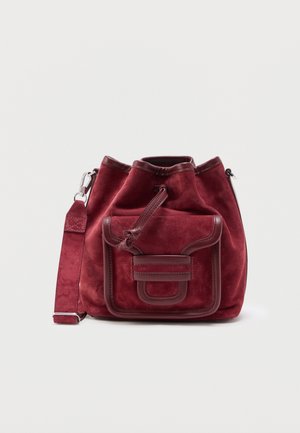 Sac seau en daim rouge avec poche à rabat frontale, garniture en cuir, fermeture à cordon et bandoulière réglable sur fond blanc.