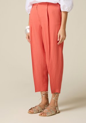 Donna con pantaloni corallo corti, camicetta bianca, bracciale d'argento e sandali beige intrecciati con lacci su sfondo semplice.