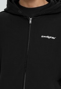 Zwarte zip-up hoodie van zachte stof, met een voorkant ritssluiting, afgewerkte manchetten en een klein wit logo op de linkerborst.