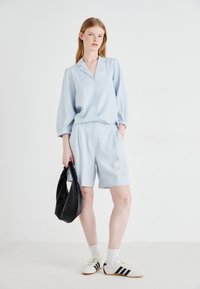 Camicia in lino azzurro chiaro con chiusura a bottoni, maniche arrotolate a tre quarti, abbinata a pantaloni corti coordinati e una borsa a tracolla nera. Sneakers bianche con strisce nere.