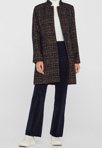 Abrigo de tweed en negro con acentos dorados y blancos, corte recto, con abertura frontal, combinado con pantalones azul marino y zapatillas blancas.