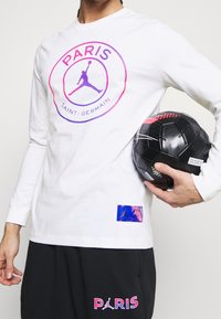 Vit långärmad skjorta med en rosa och lila Paris Saint-Germain-logga, tillsammans med svarta shorts med rosa bokstäver. Håller en svart fotboll.