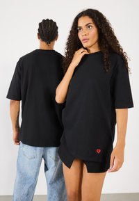 Svart oversized t-skjorter i bomull, med korte ermer og detalj av rødt hjerte-logo på kanten, vist av to modeller.