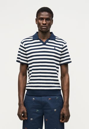 Fiatal férfi sötétkék-fehér csíkos galléros pólót és sötétkék rövidnadrágot visel, melyeken kis, sokszínű zászlóhímzés látható, egyszerű háttér előtt áll.