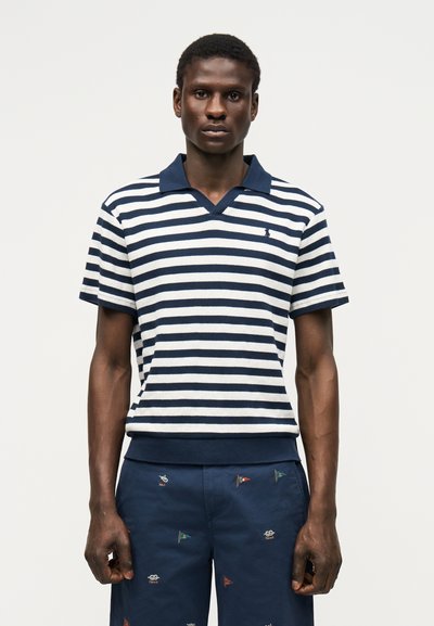 Giovane uomo che indossa una polo a righe blu navy e bianco e pantaloncini blu navy con un piccolo ricamo multicolore di una bandiera, in piedi davanti a uno sfondo semplice.