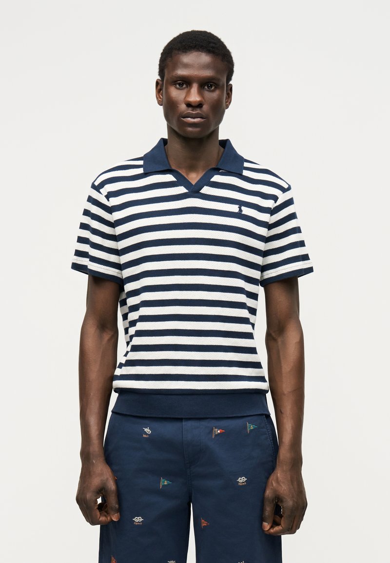 Joven con un polo a rayas azul marino y blanco y pantalones cortos azul marino con un pequeño bordado multicolor de bandera, de pie frente a un fondo liso.