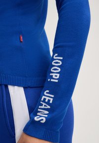Blauer Pullover mit engem Schnitt, gerippten Bündchen und weißem "JOOP! JEANS"-Text auf dem linken Arm. Das Material wirkt weich und strukturiert.