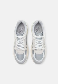 Scarpe sportive grigie e bianche che evidenziano il tessuto in rete, dettagli in pelle liscia, forma arrotondata e lacci bianchi su uno sfondo chiaro.