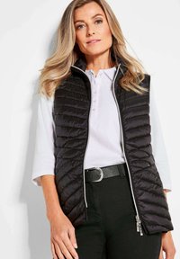 Schwarze gesteppte Pufferweste mit hohem Kragen, Frontreißverschluss und Seitentaschen, getragen über einem weißen Polohemd. Glatte Textur und figurschmeichelnde Passform.