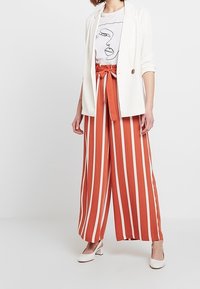 Bílý blejzr přes tričko s potiskem; volné široké kalhoty v barvě rezavé s bílými pruhy; celé doplňují bílé podpatky.