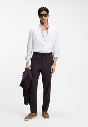 Man wearing sunglasses, white button-up shirt, black pants, brown shoes, holding a black jacket against white background.