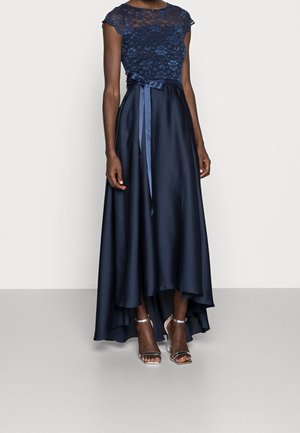 Woman wearing a navy blue high-low dress with lace top, satin skirt, and ribbon belt, paired with silver ankle-strap heels.