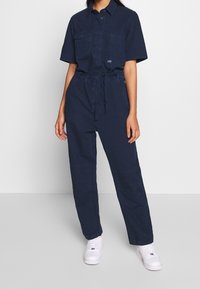 Mörkblå arbetsjumpers med korta ärmar, två framfickor, knytmidja och raka ben, tillsammans med vita sneakers.