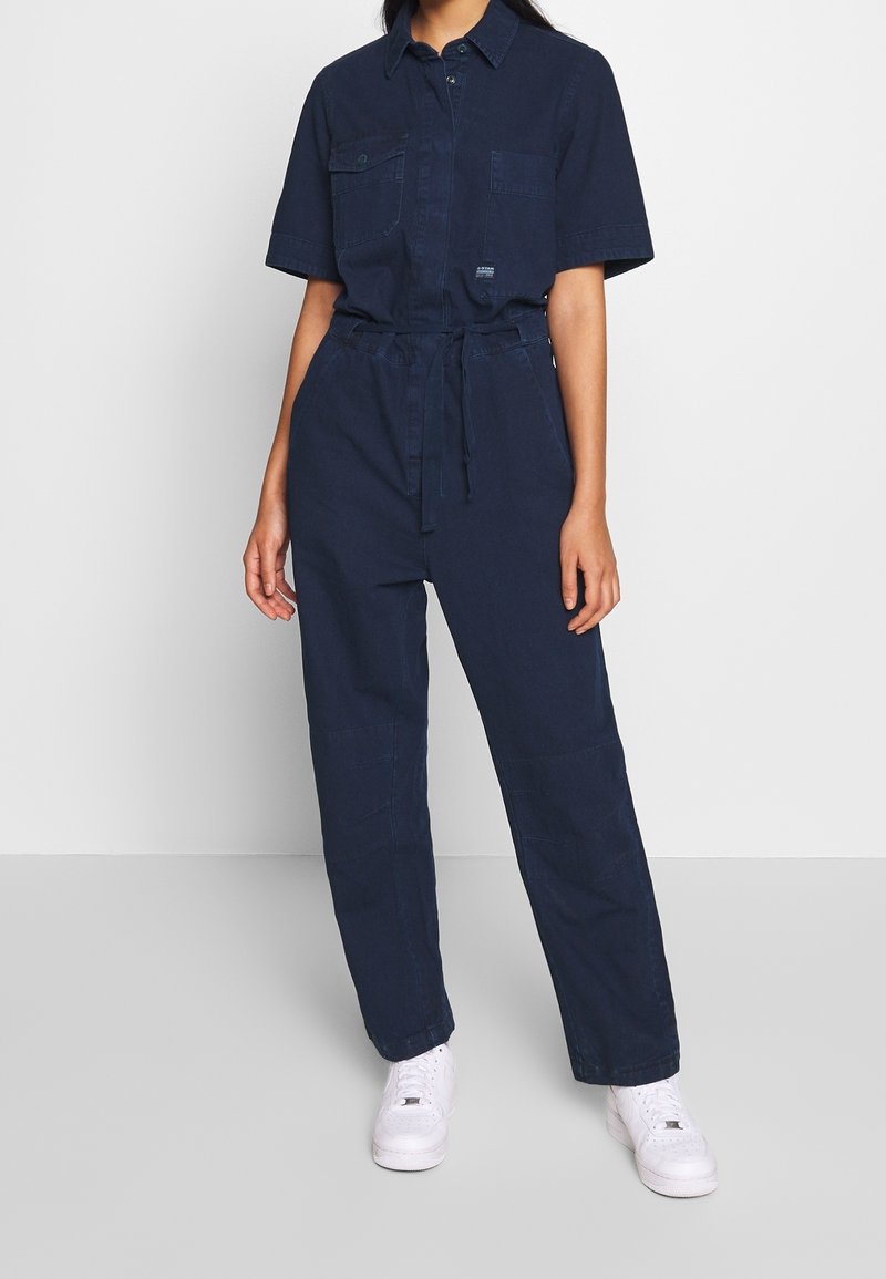 Mörkblå arbetsjumpers med korta ärmar, två framfickor, knytmidja och raka ben, tillsammans med vita sneakers.