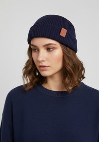 Navy blue knitted beanie with a ribbed texture and a small leather patch on the side. Worn with a navy blue sweater.