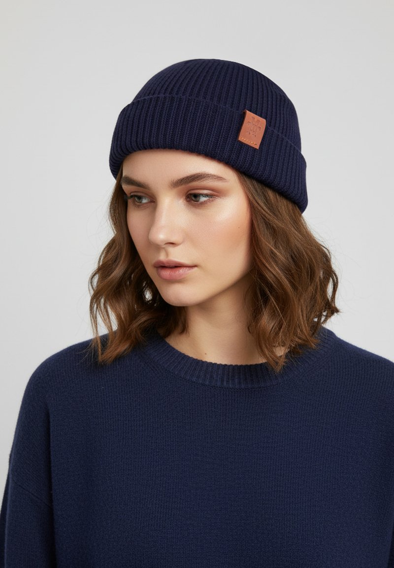 Navy blue knitted beanie with a ribbed texture and a small leather patch on the side. Worn with a navy blue sweater.