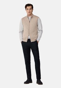 Bež prešani prsluk preko sive dukserice, u kombinaciji s crnim hlačama i tamnim cipelama. Ima prednji zip i stojeći ovratnik.