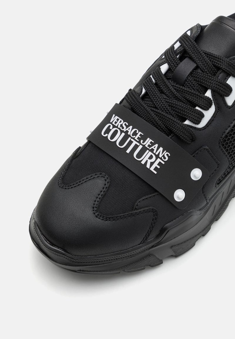 Schwarze Sneaker mit strukturierter Oberfläche, ausgestattet mit einem Netzpaneel, dicker Gummisohle und einem gebrandeten Riemen mit "VERSACE JEANS COUTURE" in Weiß.