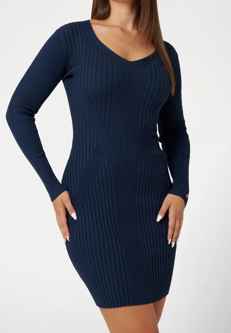 Femme portant une robe moulante en maille côtelée bleu marine à manches longues avec un col en V, les mains reposant sur les hanches.