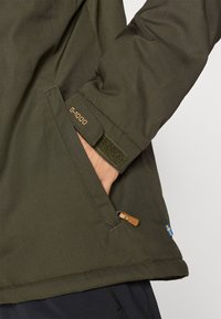 Giacca verde corta realizzata in materiale G-1000, con una tasca prominente con un accento in pelle, chiusura in velcro e dettaglio della bandiera svedese.