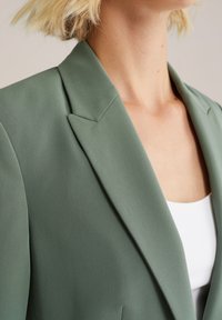 Zelený blazer se strukturovaným límcem a hladkou texturou, doplněný bílým topem pod ním. Důraz na ostré krejčovské zpracování a čisté linie.