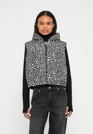 Αμάνικο γιλέκο φουσκωτό με ασπρόμαυρα στίγματα και κουκούλα, κλείσιμο με φερμουάρ και κοντό σχέδιο, φορεμένο πάνω από μαύρο μακρυμάνικο τοπ και συνδυασμένο με μαύρο τζιν.