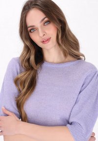 Fialový svetr s kulatým výstřihem, krátkými nabíranými rukávy a měkkým, strukturovaným pletením. Modelka pózuje se zkříženýma rukama, vlasy má vlnité.