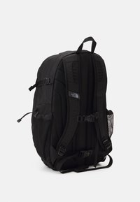 Czarny plecak North Face z wyściełanymi szelkami, zapięciem na klatce piersiowej i w pasie, bocznymi siateczkowymi kieszeniami oraz uchwytem na górze na białym tle.