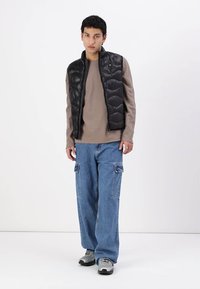 Svart polstret vest med glidelås over en taupe langermet skjorte, i kombinasjon med løse blå denim cargo bukser og grå sneakers.