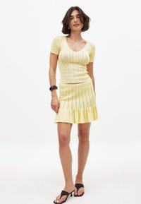 Geltonas megztas trumpomis rankovėmis viršus su V formos iškirpte ir derančia platėjančia sijona, pasižymintis griovelių tekstūra ir sluoksniuotu apvadu.