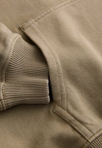 Khaki sweatshirtmaterial med en slät textur, med ribbade muddar och sömdetaljer. Designen betonar komfort och funktionalitet.