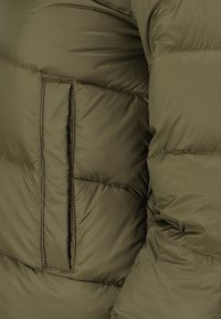 Giacca puffa verde oliva con sezioni trapuntate e una tasca laterale. Il tessuto è liscio con una texture leggermente lucida.