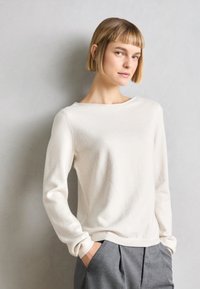 White, soft sweater with long sleeves and a boat neckline, paired with gray pleated trousers; smooth texture and minimal design.