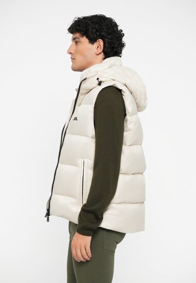 Beige puffer vest with removable hood, zip closure, and side pockets. Made from shiny, quilted material; worn over a dark green sweater.