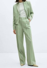 Lichtgroen op maat gemaakte pak met een fitted blazer met één zak, wijde broekspijpen en een witte top eronder. Puntige nude schoenen.