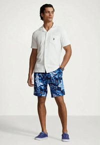 Manlig model som bär en vit kortärmad skjorta med knappar, blå blommiga shorts och blå sneakers, stående med en hand i fickan.