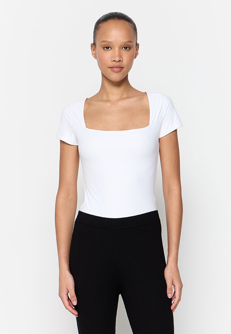 Bodysuit blanc à manches courtes avec un décolleté carré, fabriqué en tissu doux, associé à un pantalon noir à taille haute en côte.