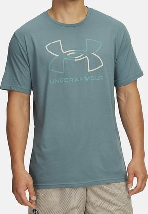 Mann trägt ein türkisfarbenes Under Armour Kurzarmshirt und hellbeige Mesh-Shorts, steht vor einem einfarbigen Hintergrund.